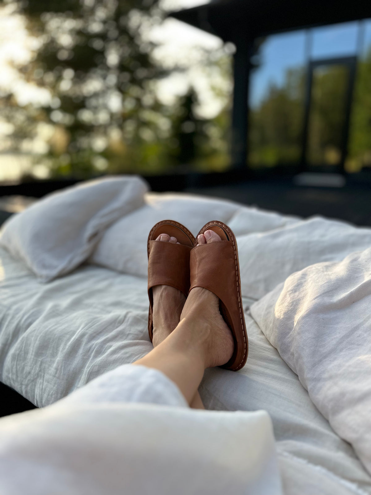 Pies con sandalias SAINT TROPEZ de cuero café sobre una cama de sábanas blancas
